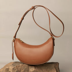 Caramel smooth leather Crescent Moon bag displayed on a textured rock surface. The bag features an adjustable long strap with gold-tone hardware and a minimalist design. Its clean, curved silhouette is accentuated by the natural background, creating a refined, elegant aesthetic.