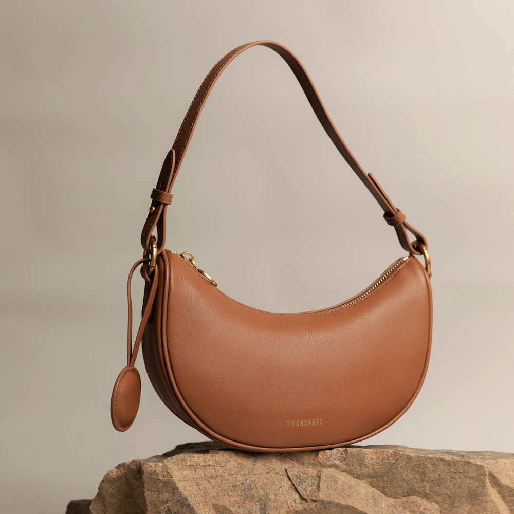Crescent Moon bag in caramel smooth leather, elegantly displayed on a stone surface with gold hardware details. The bag's curved silhouette and minimalist design are complemented by a detachable leather tag, set against a neutral-toned background, highlighting its premium craftsmanship and modern aesthetic.