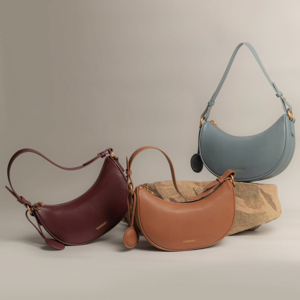 Display of three Crescent Moon bags arranged artistically on and around natural stone. From left to right, the colors showcased are wine red, caramel, and ash blue. Each bag is made from smooth leather with gold-toned hardware, accompanied by a round leather charm, and their structured silhouettes are enhanced by the soft lighting in the image