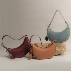 Display of three Crescent Moon bags arranged artistically on and around natural stone. From left to right, the colors showcased are wine red, caramel, and ash blue. Each bag is made from smooth leather with gold-toned hardware, accompanied by a round leather charm, and their structured silhouettes are enhanced by the soft lighting in the image