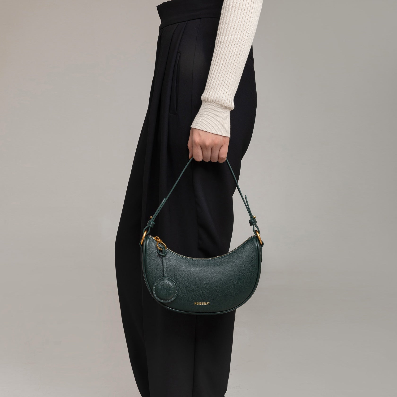 Model holding the Crescent Moon bag in pine grove smooth leather at her side, wearing a beige ribbed button-up top paired with black pleated trousers. The sleek, minimalist design of the bag is highlighted by its compact size, curved shape, and gold hardware, making it a versatile accessory for both casual and formal outfits.
