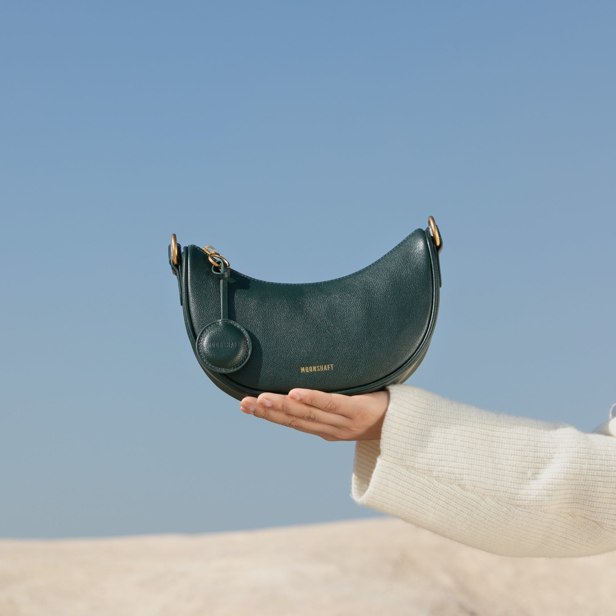 Close-up shot of a hand holding the Crescent Moon bag in pine grove green against a clear blue sky. The smooth leather bag features gold-tone hardware, a rounded silhouette, and a minimalist design with the MOONSHAFT logo in gold at the bottom. The coin pouch accessory hangs from the bag, adding a stylish detail. The backdrop of the natural landscape with soft, sandy tones enhances the elegance of the rich green leather.
