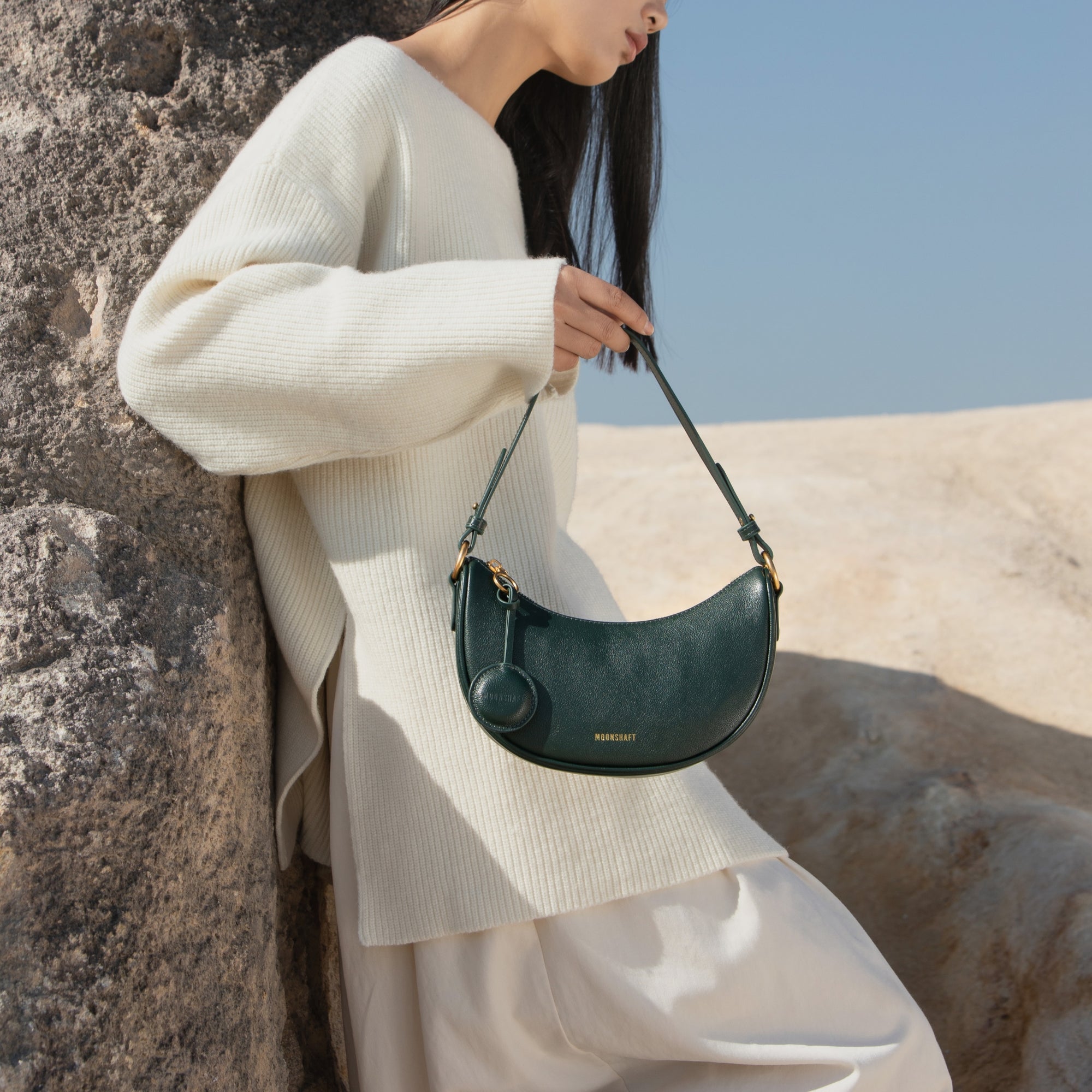 Model leaning against a textured rock in an outdoor setting, holding the Crescent Moon bag in pine grove green. The model is dressed in a soft, oversized cream sweater and light-colored pants, complementing the natural and earthy tones of the setting. The smooth leather bag, with its gold-toned hardware and minimalist design, stands out against the serene, neutral background, adding an element of sophistication to the relaxed, natural aesthetic.