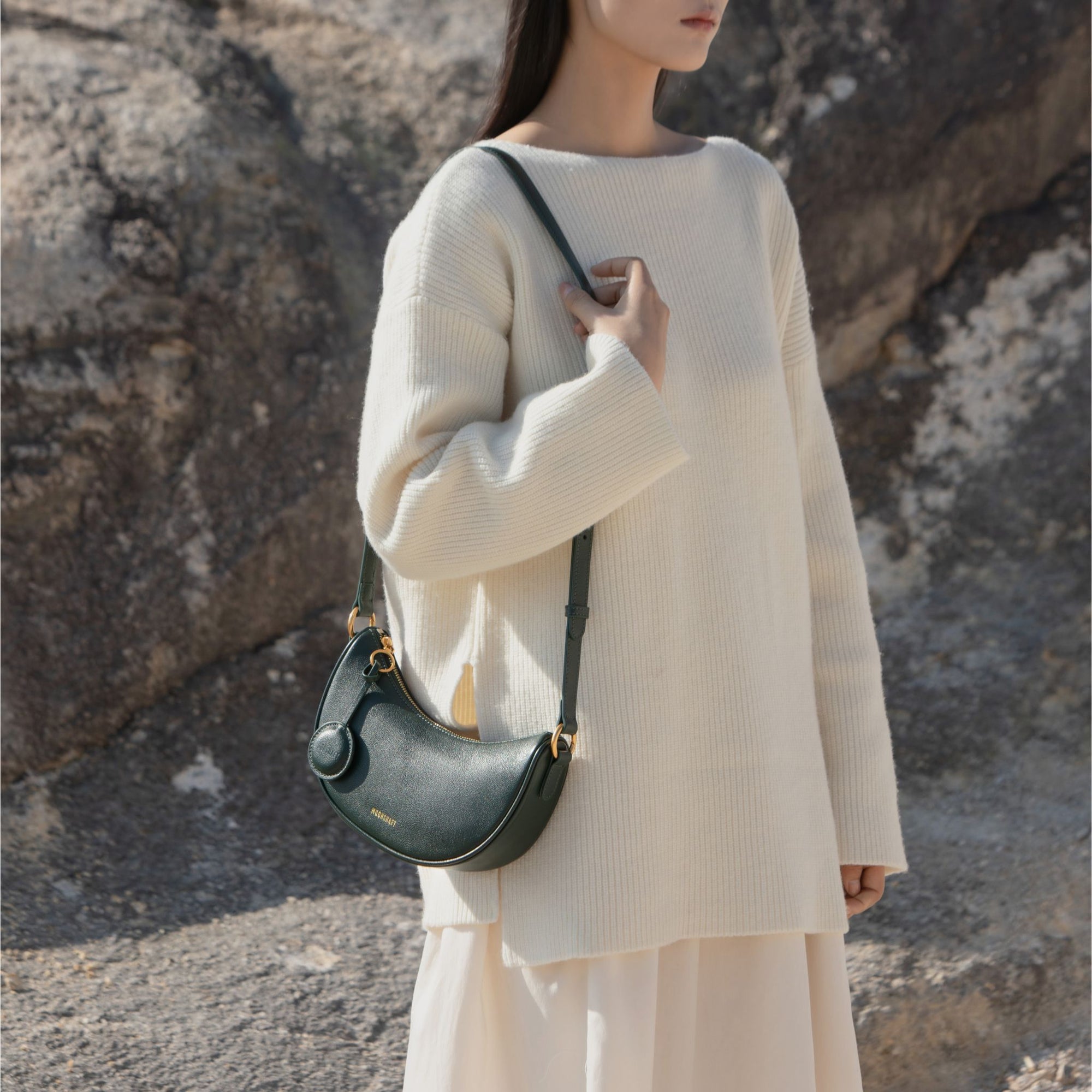 Model standing in front of a rocky background, wearing an oversized cream sweater and flowing skirt. She holds the Crescent Moon bag in pine grove green over her shoulder. The smooth leather bag features gold hardware, a rounded silhouette, and a minimalist design. The rich green color of the bag contrasts beautifully against the soft, neutral tones of the model's outfit, creating a chic and natural look.