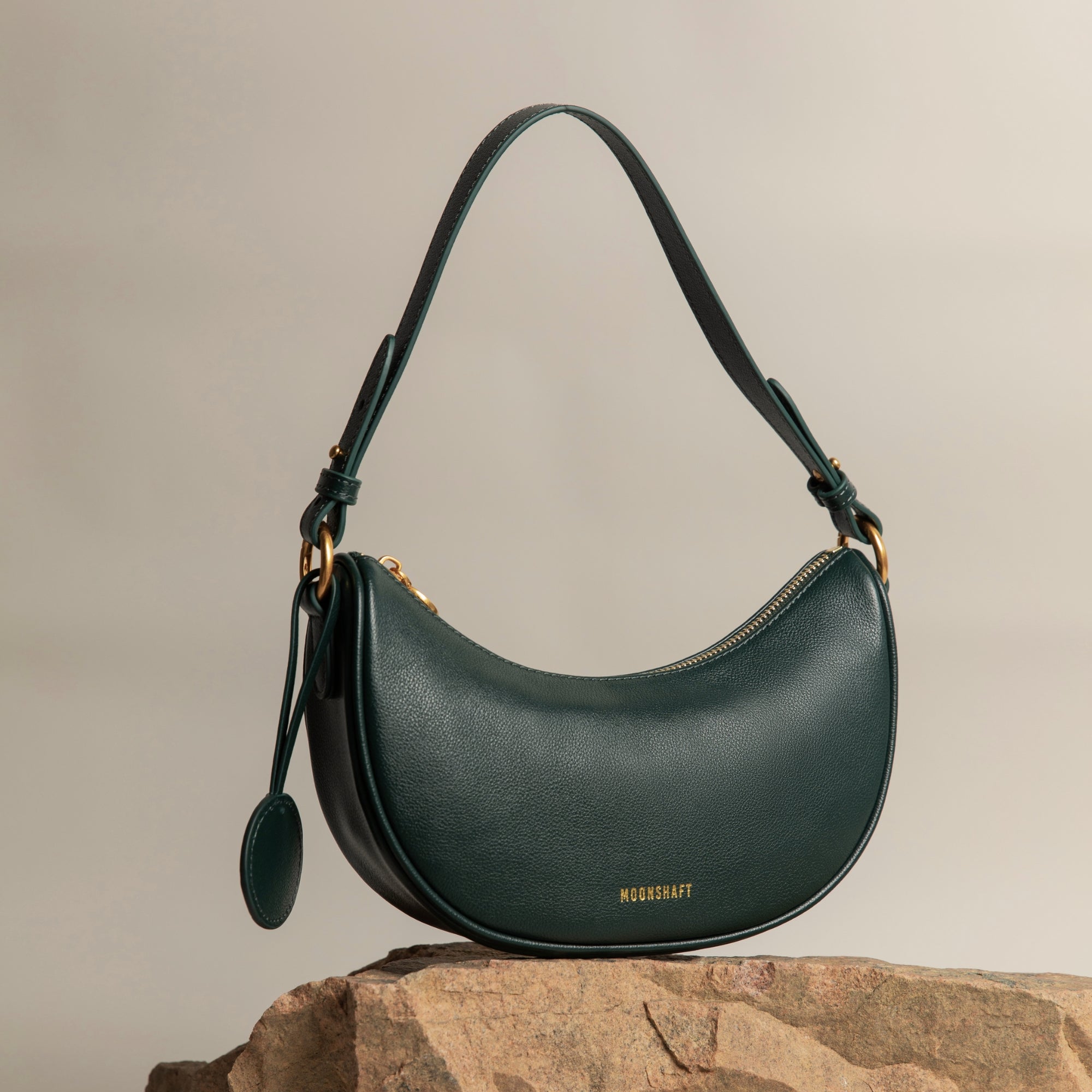 Crescent Moon bag in a deep pine grove green color, resting elegantly on a textured stone. The bag features smooth leather, gold-toned hardware, and a round leather charm hanging from the side. Its curved shape and minimalist design are complemented by the soft neutral background, highlighting the rich, luxurious tone of the bag.