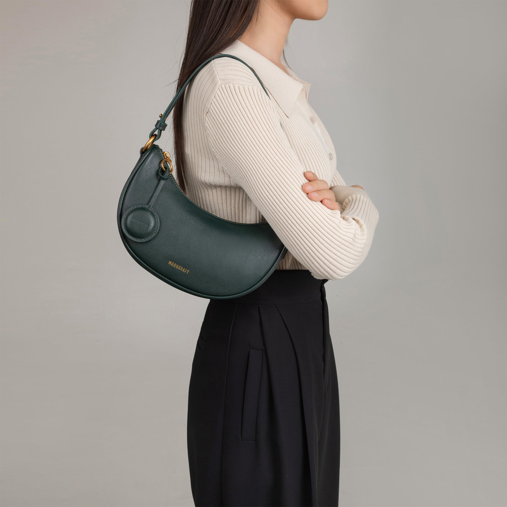 Model wearing the Crescent Moon bag in pine grove smooth leather over her shoulder. The bag is styled with a beige ribbed top and black pleated trousers, showing its versatility and chic design. The sleek, curved silhouette of the bag sits comfortably under the arm, complemented by its minimalistic details and gold hardware.