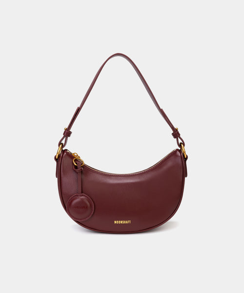 Front view of the Crescent Moon bag in wine, showcasing its smooth leather finish, curved silhouette, and gold hardware. The top handle is visible, along with an attached round coin purse accessory on the side. The minimal and elegant design highlights the bag's clean lines and structured shape.
