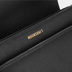 The close-up shot highlights the interior detailing of the black half-moon crossbody bag, featuring the gold-embossed MOONSHAFT logo against the smooth black leather surface. The clean stitching emphasizes the quality craftsmanship of the bag's structure.