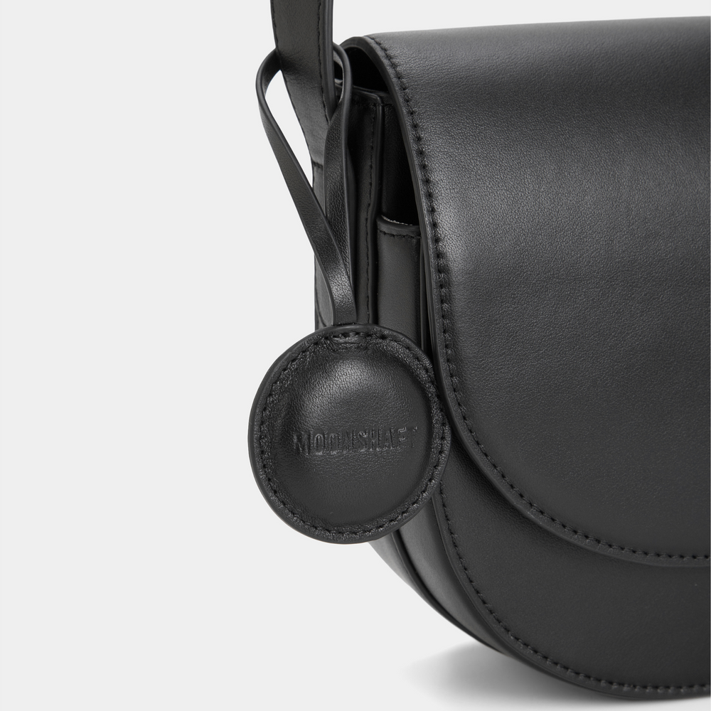 A close-up view highlighting the circular leather tag attached to the black half-moon crossbody bag. The tag features the brand's embossed logo and is positioned next to the smooth leather flap closure. The stitching and sleek design of the bag are clearly visible, emphasizing its refined craftsmanship.