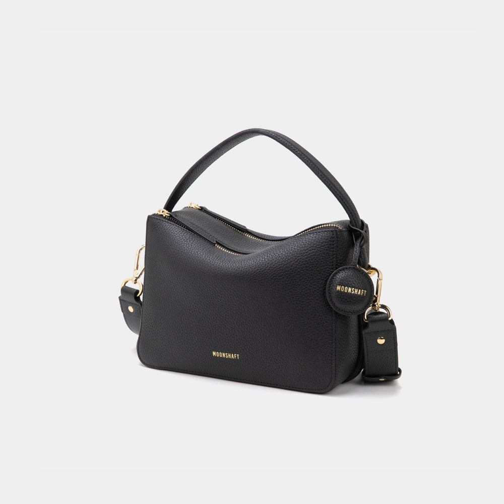 An upgraded double zip leather bag in a black color displayed against a plain background. The bag features a structured design with two visible zip compartments, showcasing its functionality and sleek style. The pebbled leather texture and subtle gold-tone zipper accents add a touch of elegance to the minimalist look. The sandstone color, a soft and neutral shade, enhances the bag's versatility, making it suitable for both casual and professional settings.