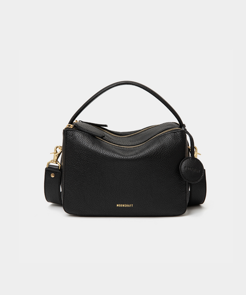 A front view of the MOONSHAFT double zip pebbled leather bag in black, displayed against a white background. The bag features a structured design with two gold-tone zippers and hardware, including a detachable strap and a top handle for versatile carrying options. The pebbled leather texture is prominent, with the MOONSHAFT logo embossed in gold on the front. A round leather charm with the brand’s logo is attached to the side, adding a stylish touch to the minimalist design.