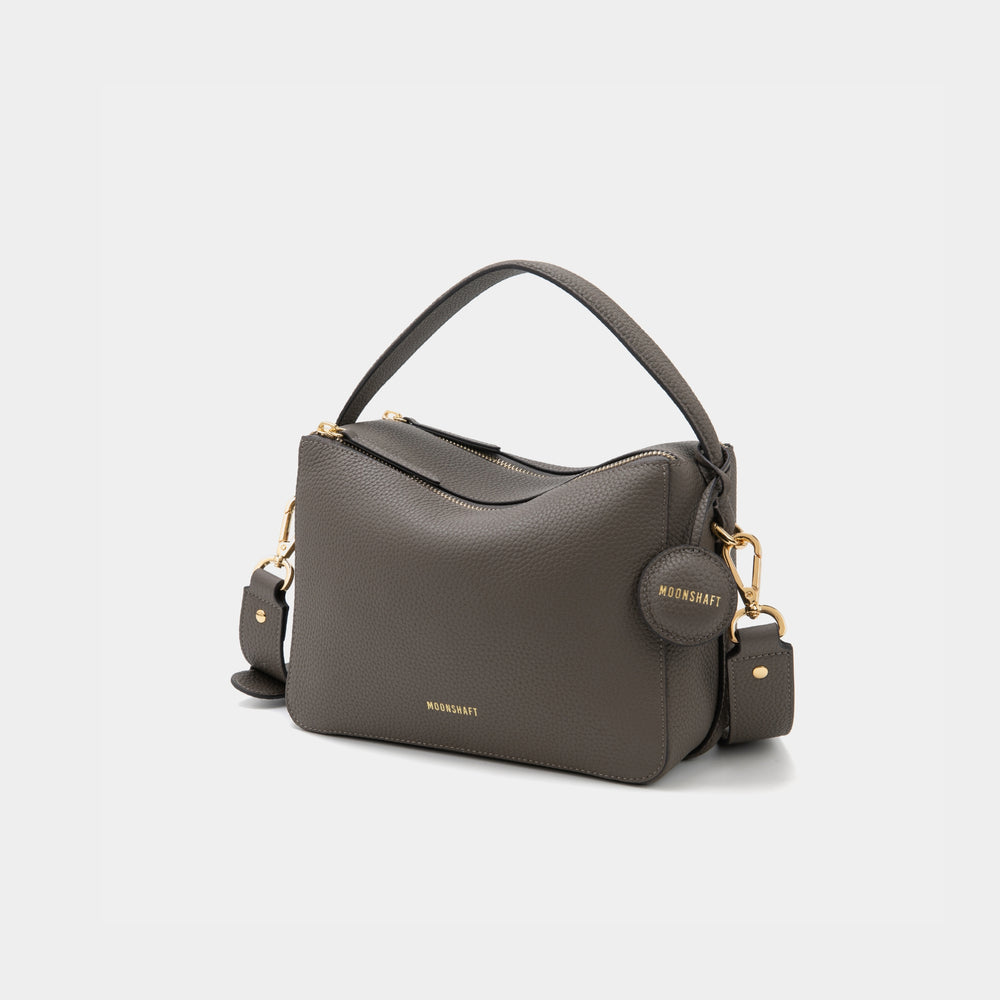 An upgraded double zip leather bag in a charcoal color displayed against a plain background. The bag features a structured design with two visible zip compartments, showcasing its functionality and sleek style. The pebbled leather texture and subtle gold-tone zipper accents add a touch of elegance to the minimalist look. The sandstone color, a soft and neutral shade, enhances the bag's versatility, making it suitable for both casual and professional settings.