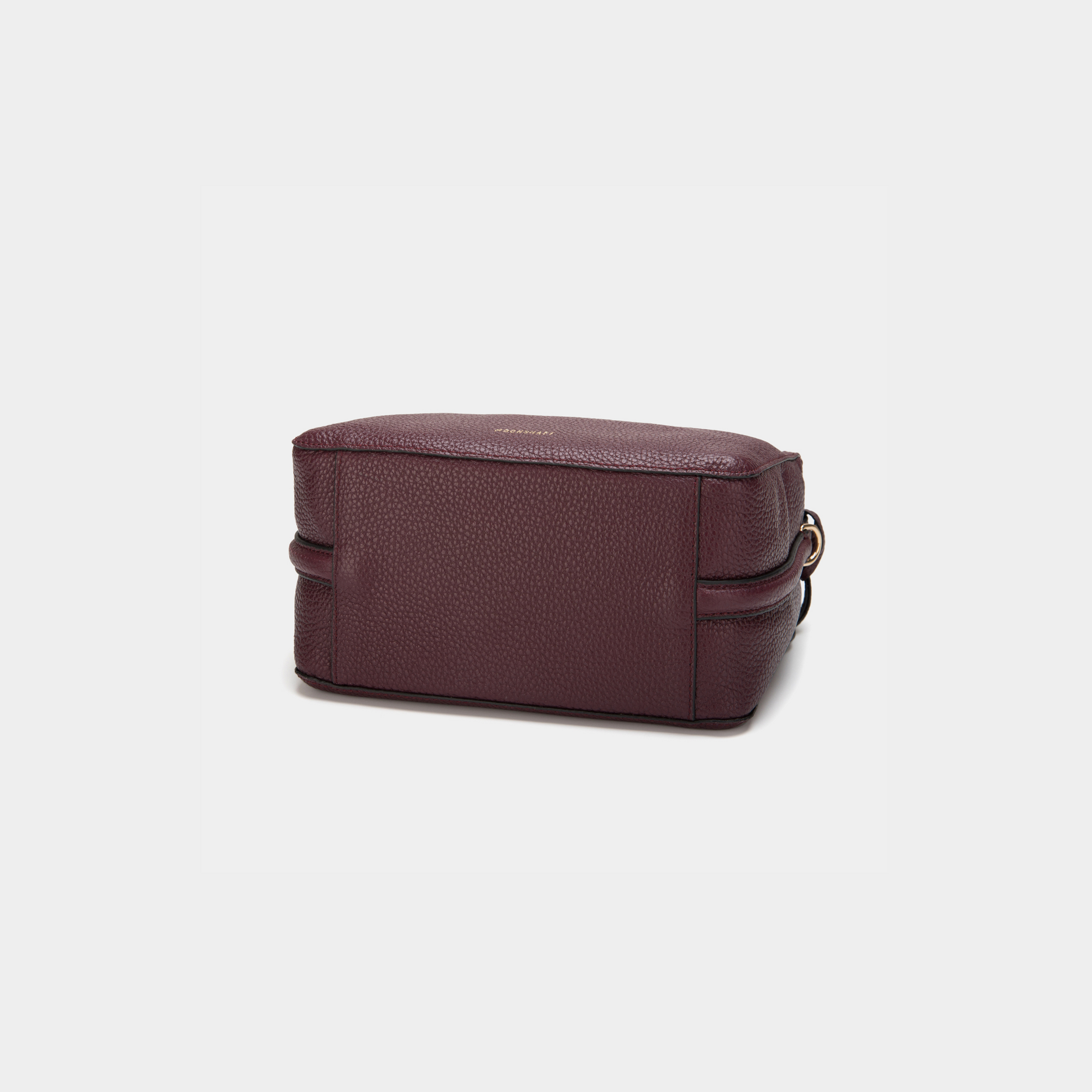 A bottom view of the MOONSHAFT double zip pebbled leather bag in wine, highlighting the structured rectangular base with clean stitching along the edges. The smooth pebbled leather texture is prominent, and the side profile reveals the compact, sleek design of the bag. A glimpse of the attached round leather charm is visible to the side.