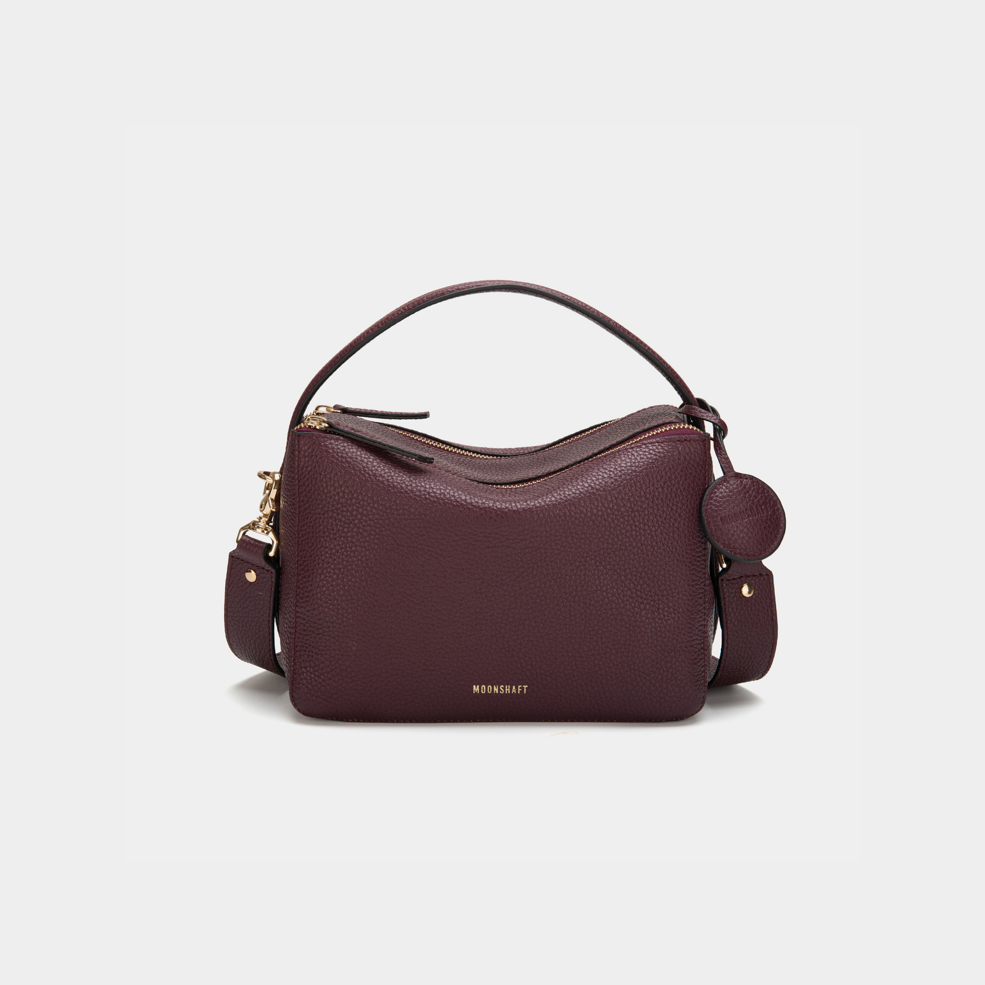 A front view of the MOONSHAFT double zip pebbled leather bag in wine, displayed against a white background. The bag features a structured design with two gold-tone zippers and hardware, including a detachable strap and a top handle for versatile carrying options. The pebbled leather texture is prominent, with the MOONSHAFT logo embossed in gold on the front. A round leather charm with the brand’s logo is attached to the side, adding a stylish touch to the minimalist design.