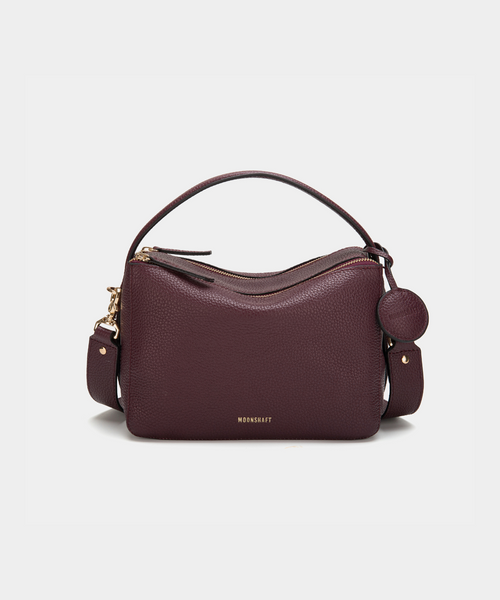 A front view of the MOONSHAFT double zip pebbled leather bag in wine, displayed against a white background. The bag features a structured design with two gold-tone zippers and hardware, including a detachable strap and a top handle for versatile carrying options. The pebbled leather texture is prominent, with the MOONSHAFT logo embossed in gold on the front. A round leather charm with the brand’s logo is attached to the side, adding a stylish touch to the minimalist design.