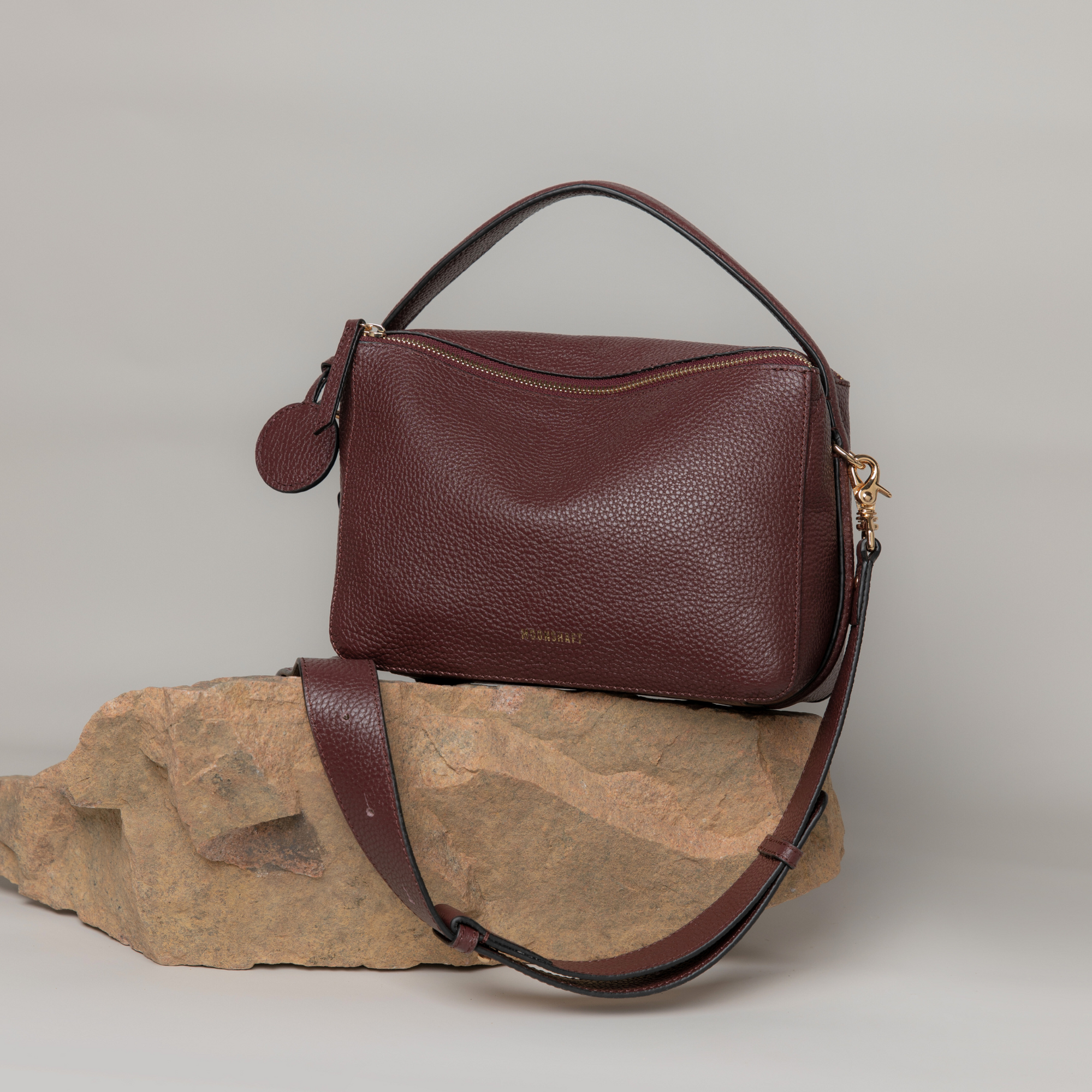Wine MOONSHAFT double zip pebbled leather bag displayed on a natural stone surface against a neutral background. The bag features gold-tone zippers, a top handle, and a detachable strap. The rich pebbled leather texture stands out, with a round leather charm attached to the side. The earthy stone element adds a rugged contrast to the bag's sleek and elegant design.