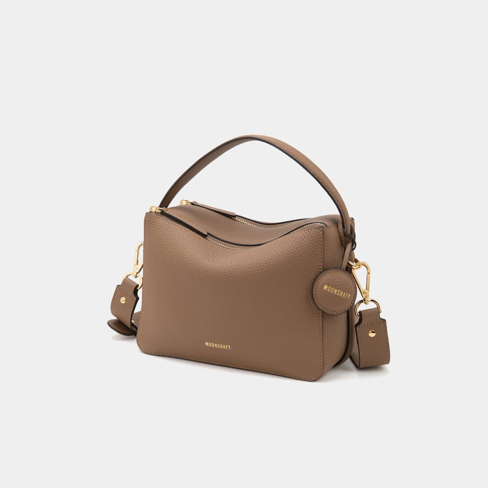 An upgraded double zip leather bag in a driftwood color displayed against a plain background. The bag features a structured design with two visible zip compartments, showcasing its functionality and sleek style. The smooth leather texture and subtle gold-tone zipper accents add a touch of elegance to the minimalist look. The sandstone color, a soft and neutral shade, enhances the bag's versatility, making it suitable for both casual and professional settings.