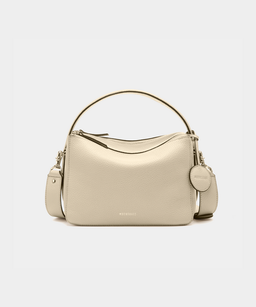 A front view of the MOONSHAFT double zip pebbled leather bag in sand, displayed against a white background. The bag features a structured design with two gold-tone zippers and hardware, including a detachable strap and a top handle for versatile carrying options. The pebbled leather texture is prominent, with the MOONSHAFT logo embossed in gold on the front. A round leather charm with the brand’s logo is attached to the side, adding a stylish touch to the minimalist design.