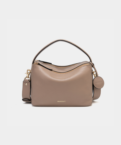 Front view of the MOONSHAFT double zip pebbled leather bag in taupe, featuring gold-tone zippers, a top handle, and a detachable strap. The MOONSHAFT logo is embossed in gold on the front, with a round leather charm attached.