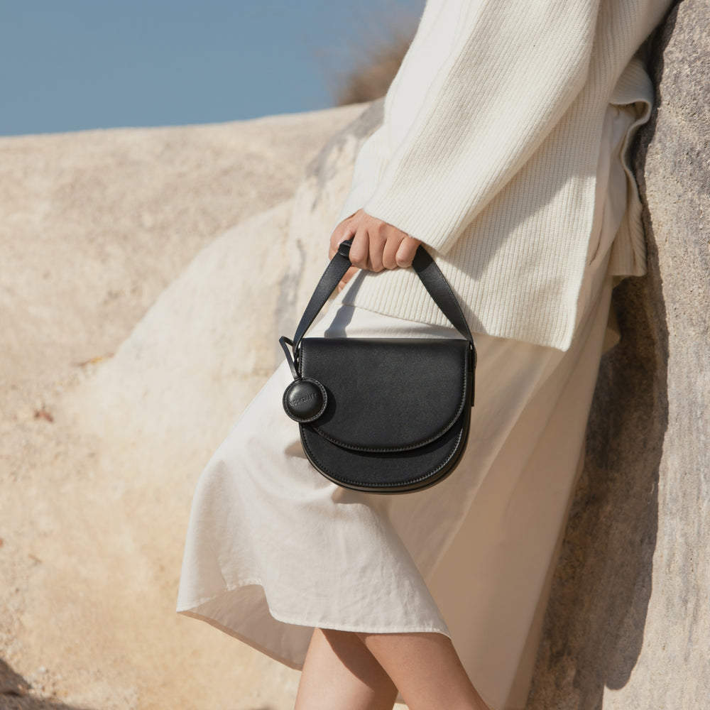A model leans casually against a rock, holding the black half-moon crossbody bag by its leather strap. The bag's structured silhouette contrasts with the soft, flowing cream sweater and skirt. The circular leather tag attached to the bag adds a subtle detail to the minimalistic design, while the natural rocky backdrop highlights the bag's sleek appearance. The relaxed posture and neutral tones create a harmonious and effortless look.