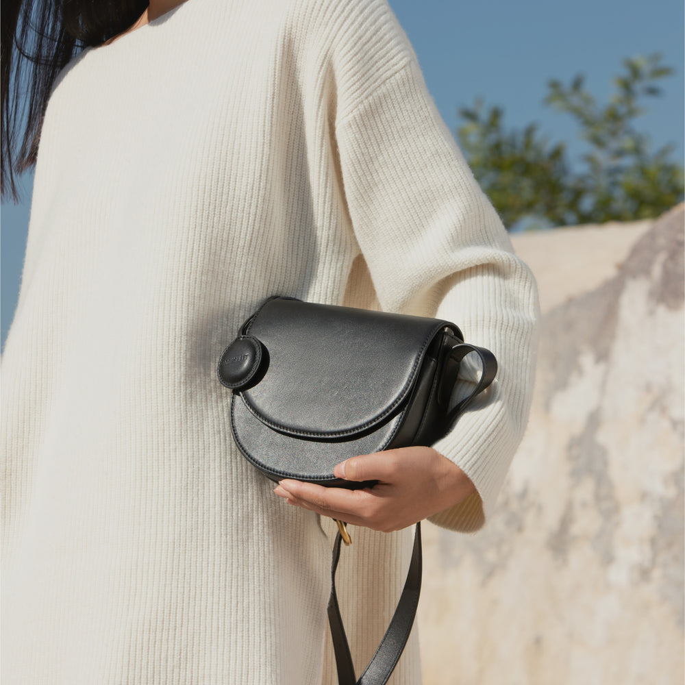 A model, dressed in a soft cream sweater, holds the half-moon crossbody bag in black by her side. The sleek and smooth leather texture of the bag contrasts with the softness of her outfit. The circular leather tag adds a stylish detail to the compact, curved design of the bag. The natural background of rocks and greenery enhances the simplicity and elegance of the overall look.