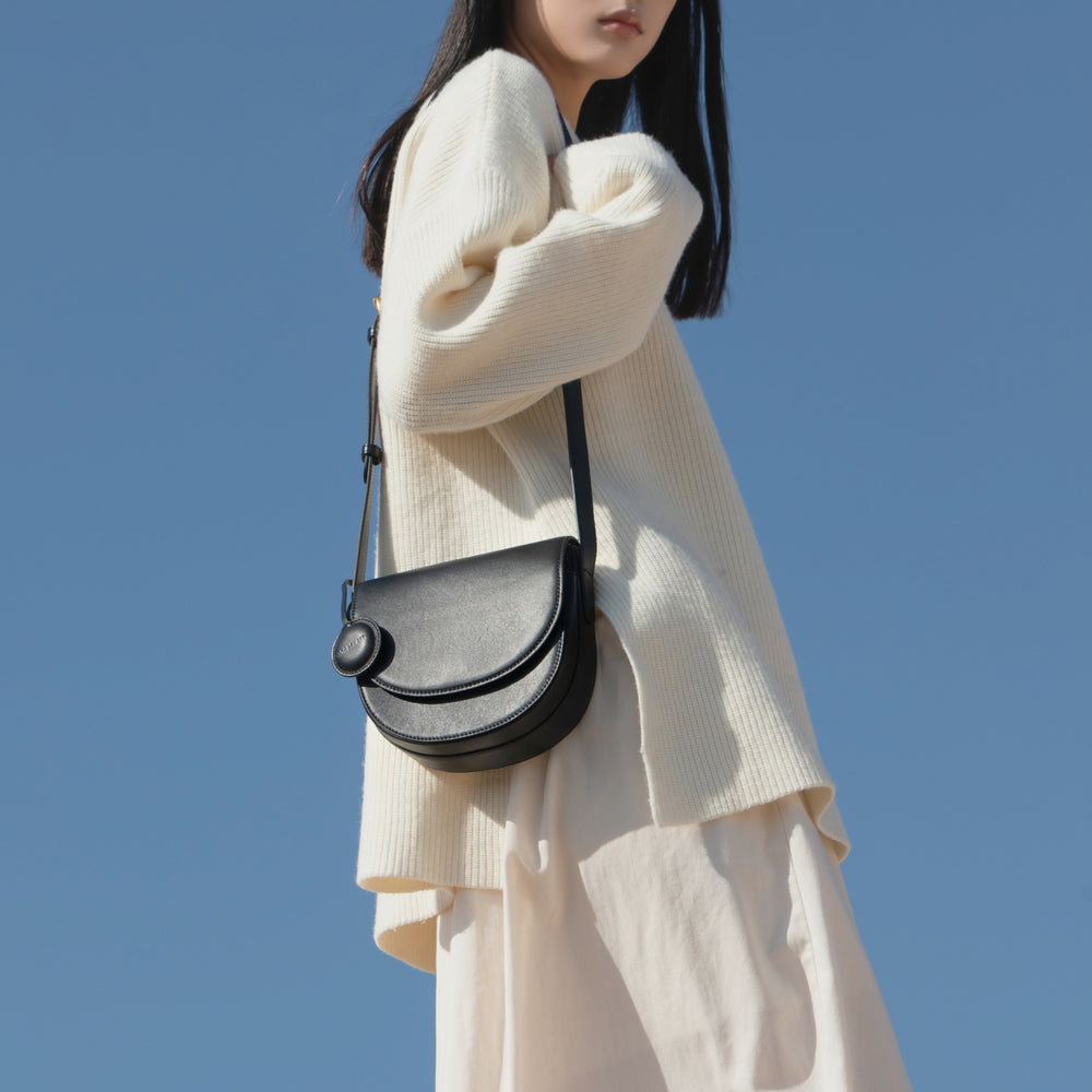 A model wearing a loose, cream-colored sweater paired with a flowing skirt, styled with a black half-moon crossbody bag. The smooth leather bag features a curved, structured design and a circular leather tag. The bag's minimalist elegance contrasts against the soft, neutral outfit, creating a chic and sophisticated look against the backdrop of a clear blue sky.