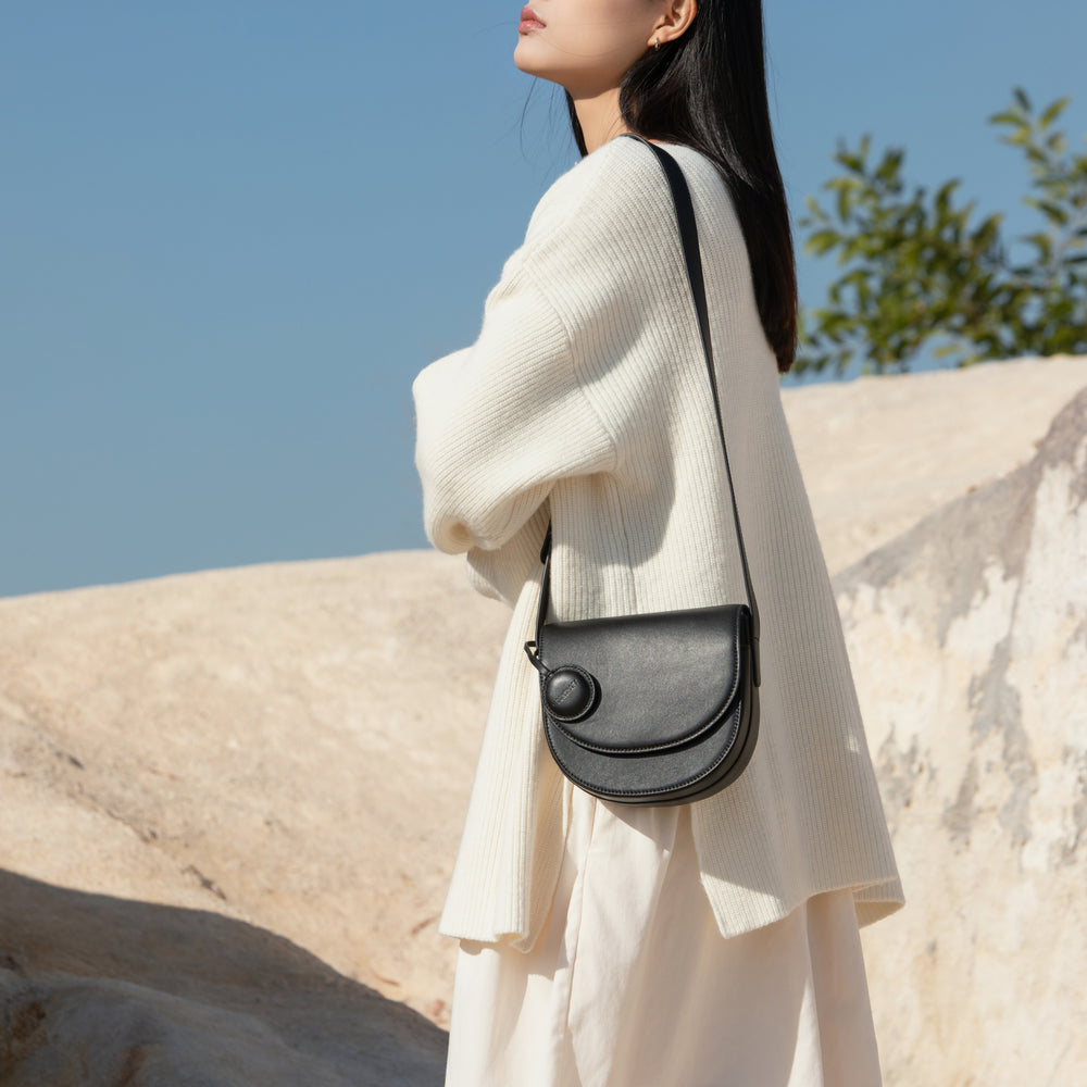 A model is wearing a soft oversized cream sweater and a flowing skirt while carrying the half-moon crossbody bag in black. The bag hangs comfortably across her body, showcasing the sleek leather finish and the circular leather tag. The setting features natural light against a backdrop of smooth rocks and trees, highlighting the effortless, minimalistic design of the bag in a chic outdoor environment.