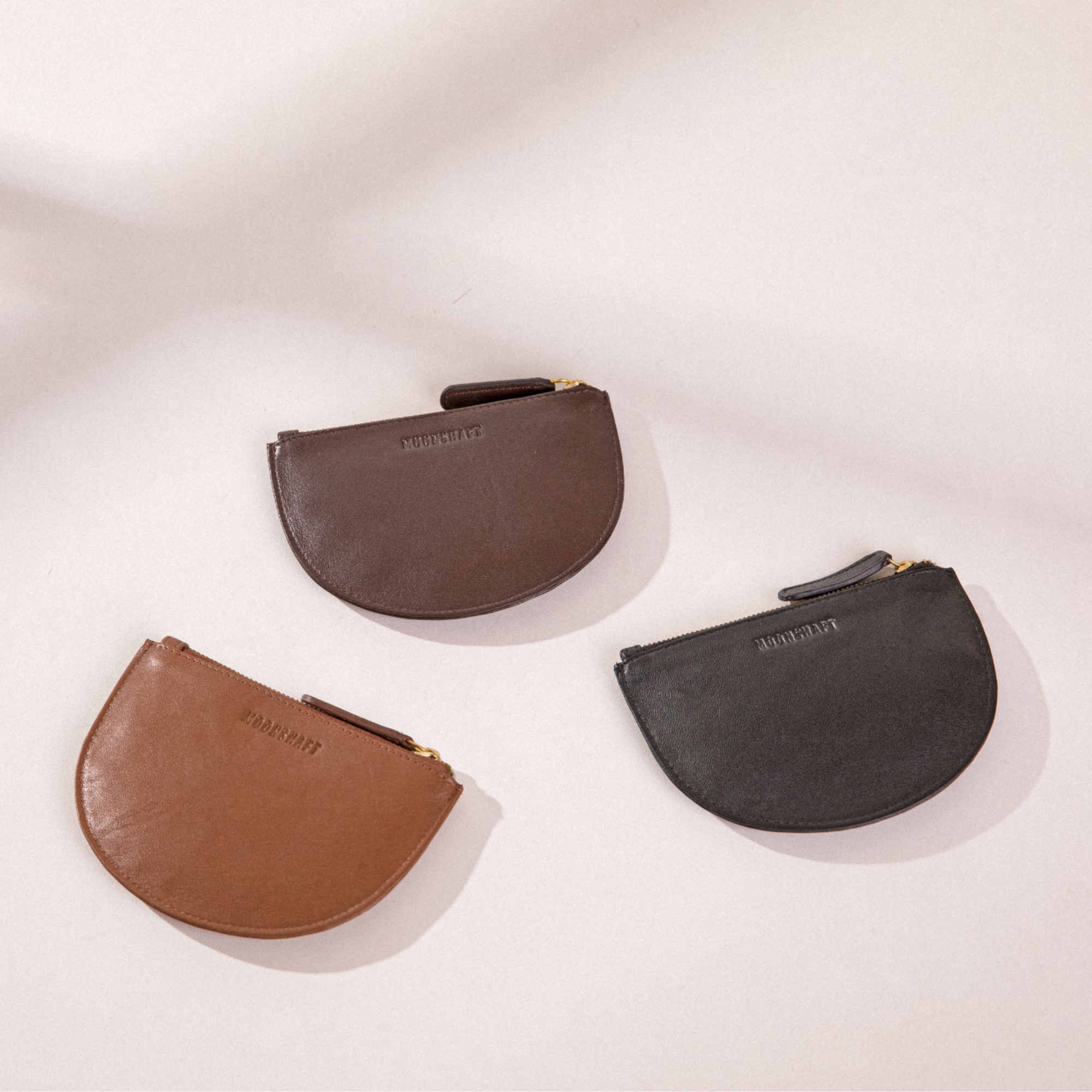  The image shows three Half Moon Coin Purses made of goat leather, arranged on a light background. The purses are in different colors: one in cognac, one in brown, and one in black. Each purse features the MOONSHAFT logo embossed on the front and is equipped with a gold-tone zipper with a matching leather pull. Shadows from the purses add dimension to the composition, highlighting their curved shape.