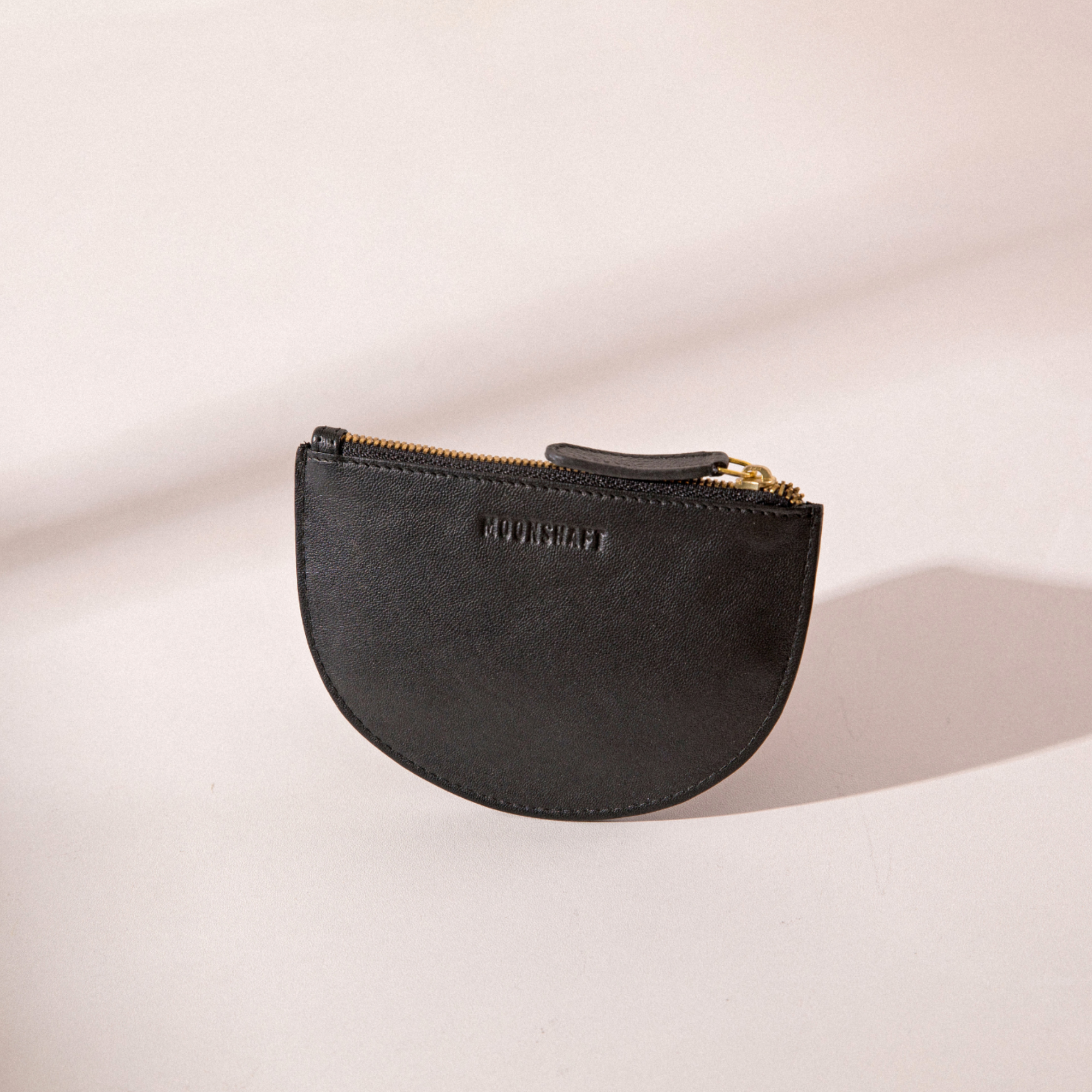 The image features the Half Moon Coin Purse in black goat leather, standing upright against a light background. The MOONSHAFT logo is embossed on the front, and the purse is equipped with a gold-tone zipper along the top, complemented by a matching black leather pull. Gentle shadows cast from the product emphasize its sleek design and soft leather texture.