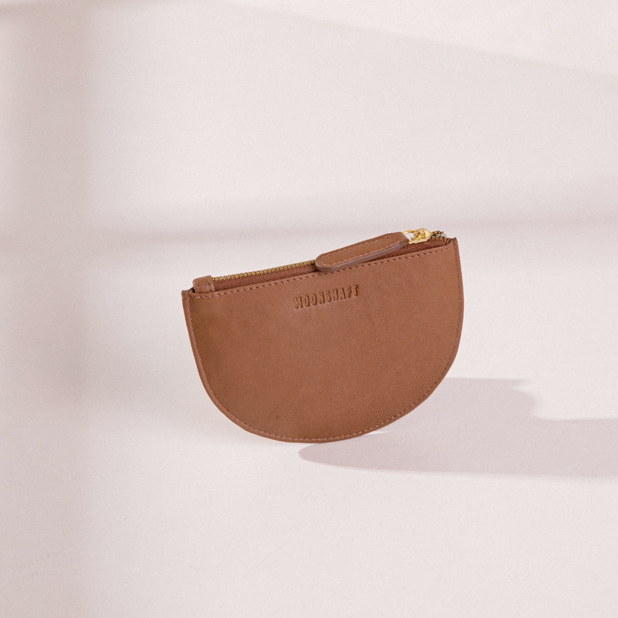The Half Moon Coin Purse in cognac goat leather, standing upright on a light background. The MOONSHAFT logo is embossed on the front of the purse, and a gold-tone zipper with a matching tan leather pull is visible at the top. Soft shadows highlight the purse's smooth leather texture and minimalist design.