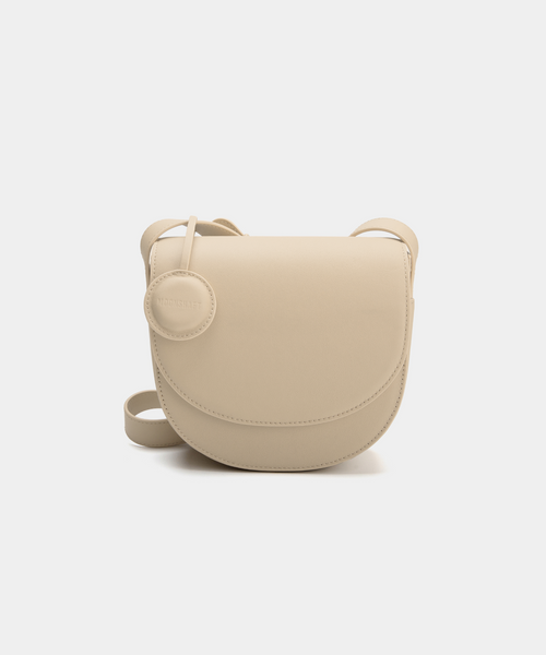 A beige half-moon crossbody bag with a smooth leather finish, featuring a circular leather tag attached to the strap. The structured design highlights the bag's minimalist aesthetic and sleek curves, offering an elegant yet functional style for everyday wear.