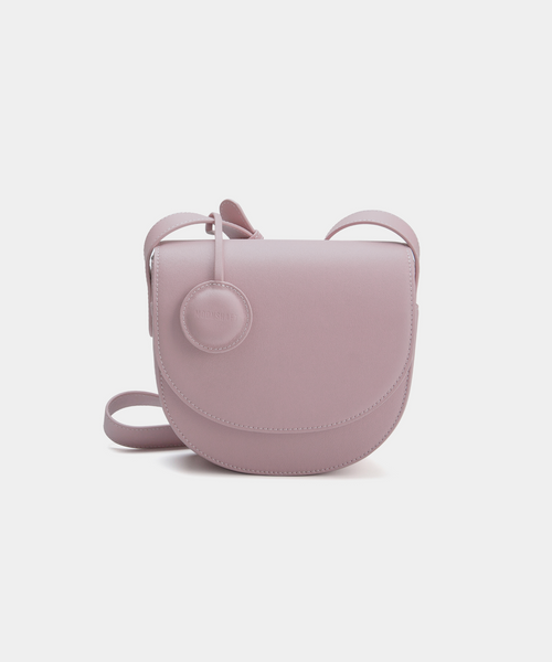 A Light Lilac half-moon crossbody bag with a sleek, smooth leather finish. It features a circular leather tag attached to the strap and a simple, structured design. The bag has a curved flap closure and an adjustable crossbody strap.