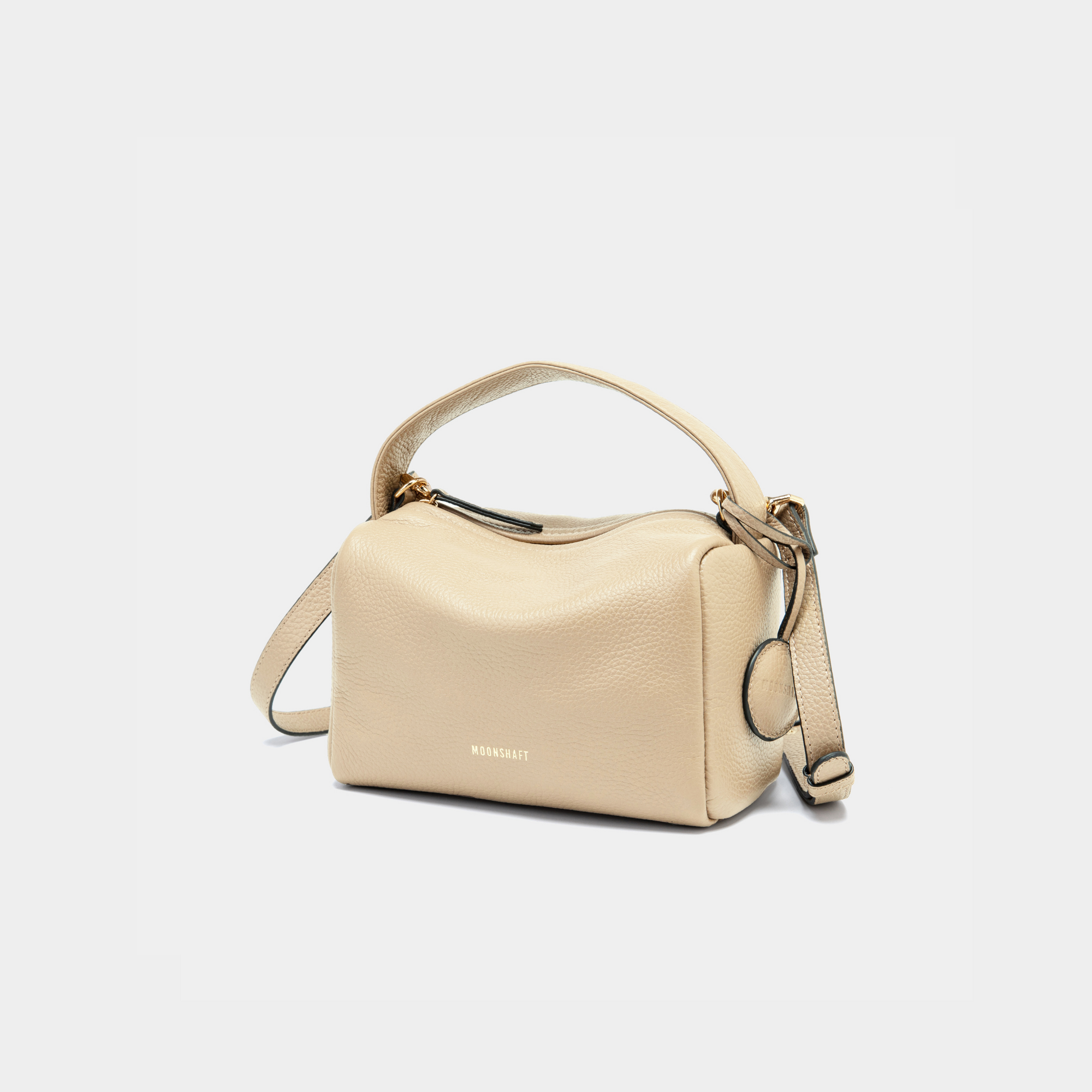 The image shows the Moonshaft Loaf Bag in sand pebbled leather, captured from a front-side angle. The bag's boxy, structured design is highlighted, along with the top handle and detachable shoulder strap draping down the side. The Moonshaft logo is clearly visible on the front.