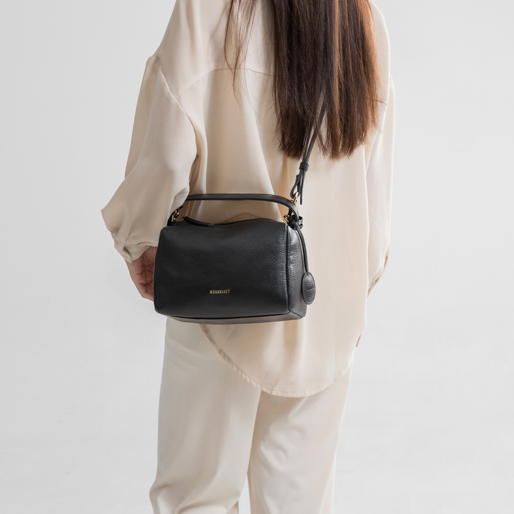 A model is showcasing the black Loaf Bag in a casual shoulder style. The bag has a boxy and structured design, made of pebbled leather, adding a luxurious texture. The gold-tone branding and hardware create a sophisticated contrast against the deep black color of the bag. The model is dressed in a soft, neutral-toned outfit, which highlights the bag's minimalist elegance and versatility. This look emphasizes how the black Loaf Bag can seamlessly complement both casual and chic ensembles.