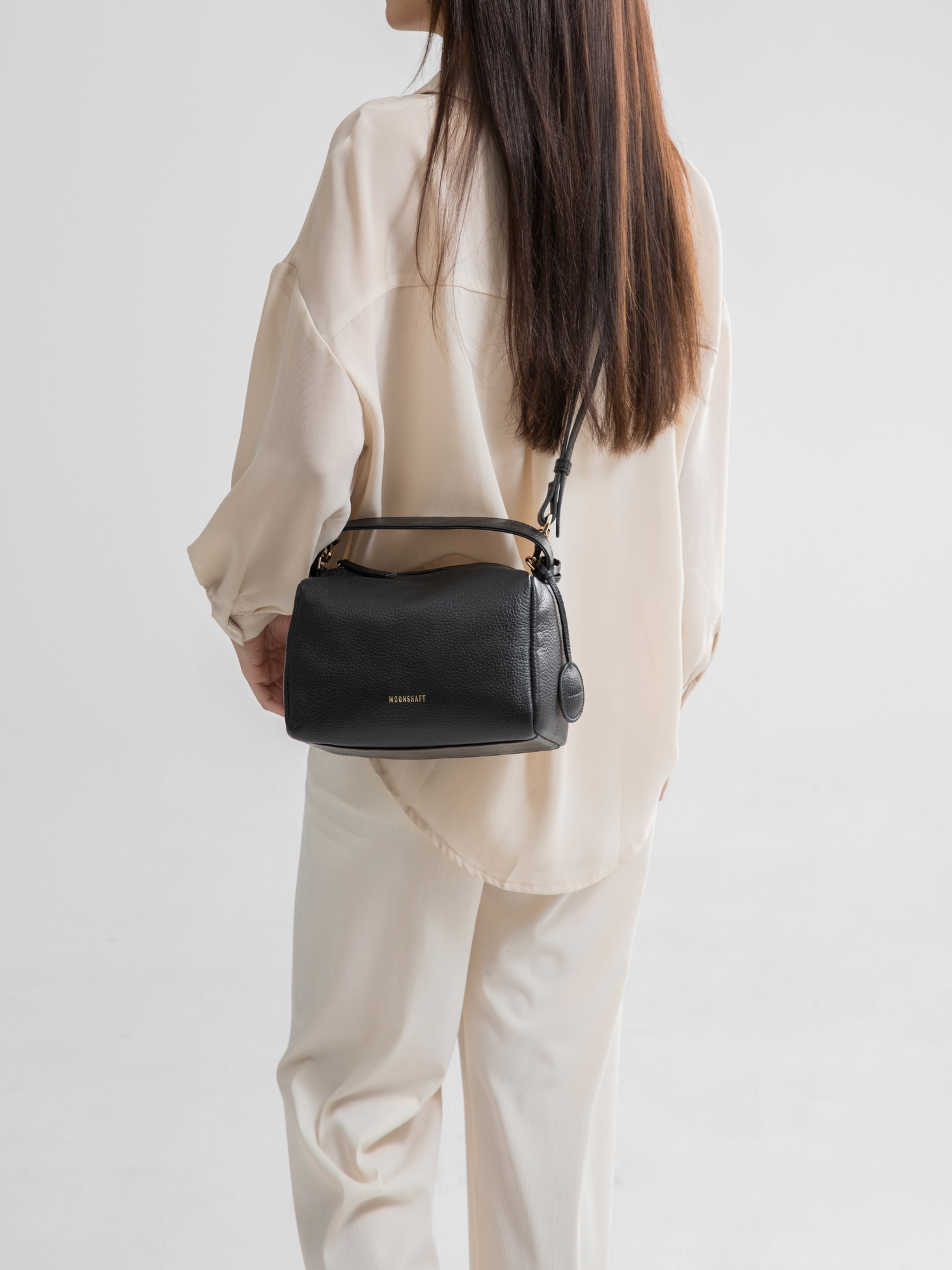 A model is showcasing the black Loaf Bag in a casual shoulder style. The bag has a boxy and structured design, made of pebbled leather, adding a luxurious texture. The gold-tone branding and hardware create a sophisticated contrast against the deep black color of the bag. The model is dressed in a soft, neutral-toned outfit, which highlights the bag's minimalist elegance and versatility. This look emphasizes how the black Loaf Bag can seamlessly complement both casual and chic ensembles.