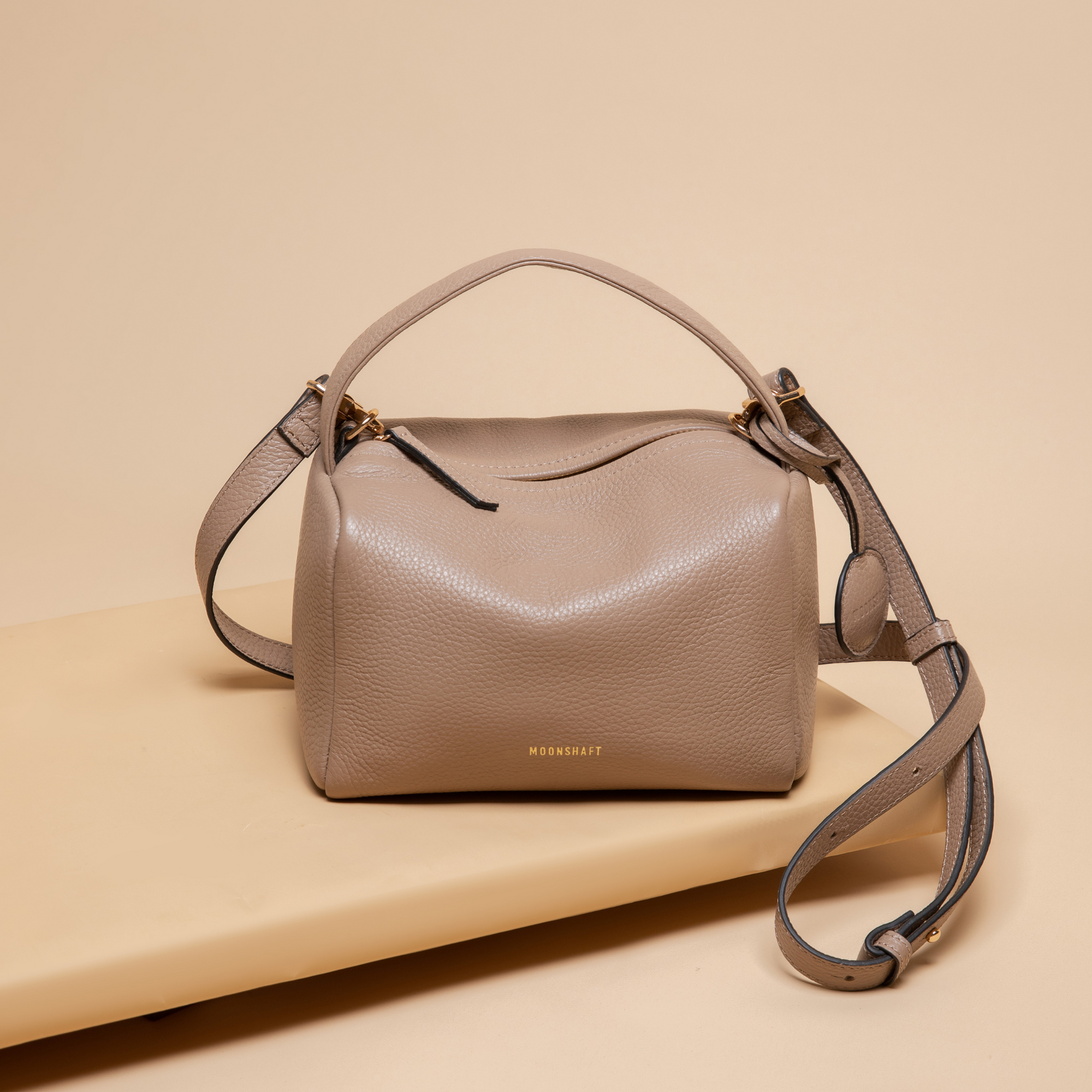 A taupe-colored Loaf Bag displayed on a beige surface, showcasing its boxy, structured design made from pebbled leather. The bag features gold-tone hardware, a top handle, and a detachable shoulder strap with adjustable length. The brand name "MOONSHAFT" is embossed in gold on the lower front, adding a touch of sophistication. The bag's clean lines and minimalist aesthetic highlight its functional yet stylish design.