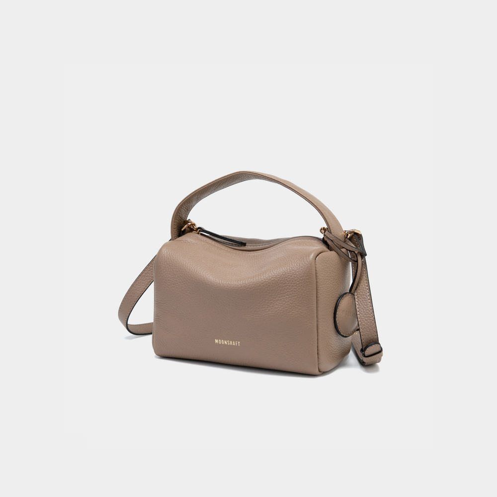 This is a three-quarter view of the Moonshaft Loaf Bag in taupe color. The image highlights the structured, boxy silhouette of the bag made from pebbled leather. The top handle is shown curving smoothly across the top, while the adjustable shoulder strap hangs on the side. The taupe leather is soft and neutral, exuding elegance and versatility. A small round leather charm is attached to the side, adding a minimalist accent. 