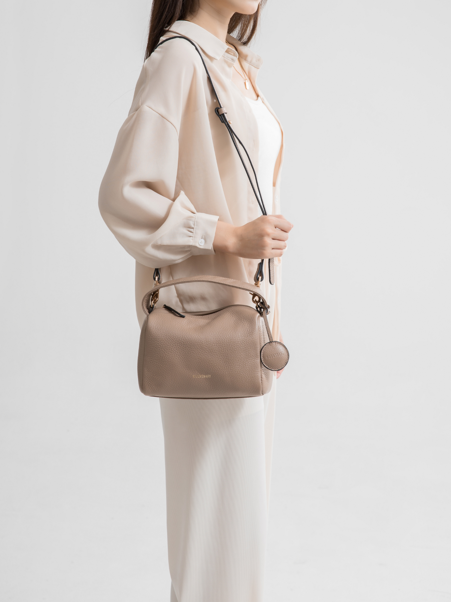 A model is seen carrying the taupe Loaf Bag with a detachable shoulder strap in a crossbody style. The bag, made of pebbled leather, features a structured and boxy design. The bag's gold zipper closure and matching gold embossed logo on the front add an elegant touch. The model is dressed in a soft, neutral-toned outfit, complementing the bag's understated and versatile color. The overall look is chic and minimalist, perfect for everyday wear.