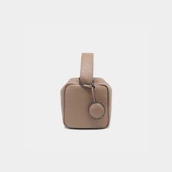 This is a side view of the Moonshaft Loaf Bag in taupe color. The structured, boxy shape is emphasized, with the pebbled leather texture clearly visible. The handle on the top is short and firm, suggesting versatility for hand carrying. A round leather charm, attached to the side of the bag, adds a subtle accent. The taupe shade provides a neutral and elegant tone, perfect for a sophisticated and minimalist style. The clean lines and well-defined edges contribute to the refined aesthetic of the bag.