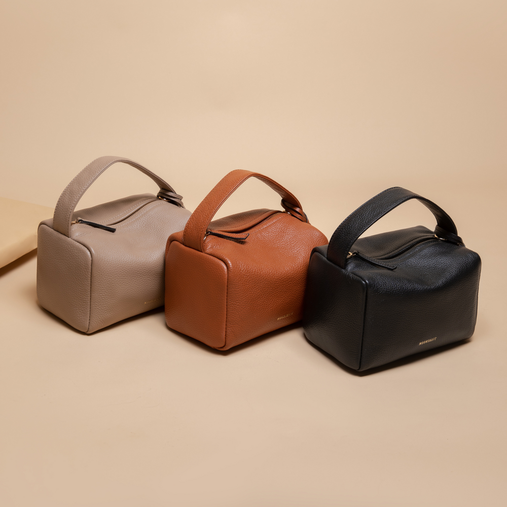 Three Loaf Bags are displayed side by side on a beige background. The bags feature a structured, rectangular silhouette made of pebbled leather. They come in three colors: taupe, tan, and black. Each bag is equipped with a top handle and a zippered closure, with gold-tone hardware accents and the brand name "MOONSHAFT" embossed in gold. The bags have a clean, minimalist design, emphasizing functionality and style.