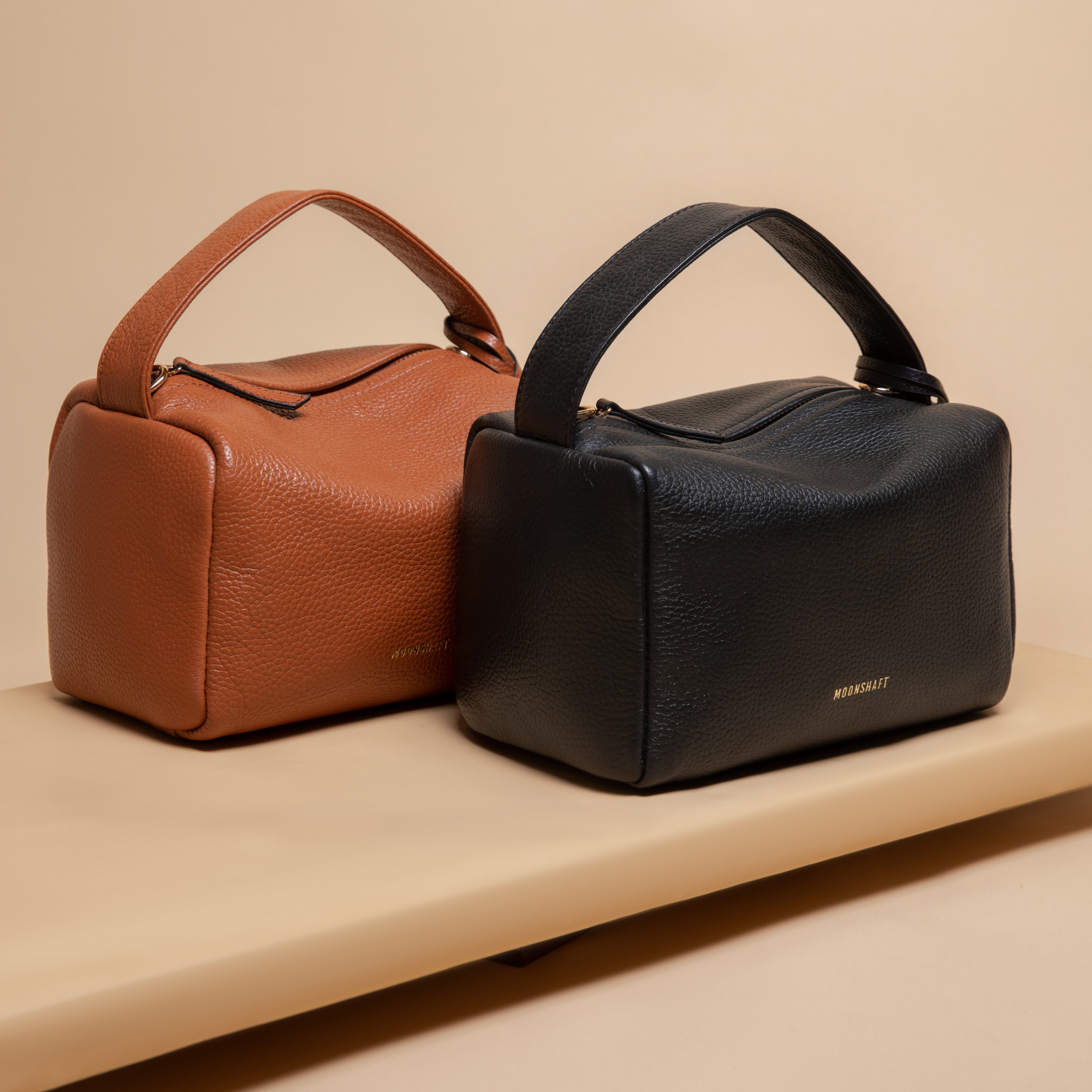 Two Loaf Bags are positioned side by side on a beige surface. The bag on the left is in a rich tan color, while the one on the right is in classic black. Both bags feature a structured design with top handles, smooth pebbled leather, and gold-tone branding, offering a sleek and elegant look.
