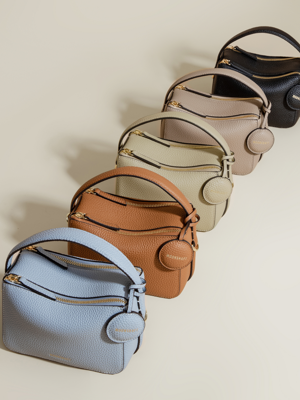 A lineup of Mini Double Zip Bags in five different colors: aquamarine, toffee, beige, taupe, and black. Each bag is made from pebbled leather, featuring a top handle, gold-tone hardware, and a round leather tag embossed with the 'MOONSHAFT' logo. The structured, minimalist design of the bags highlights their modern and versatile appeal.