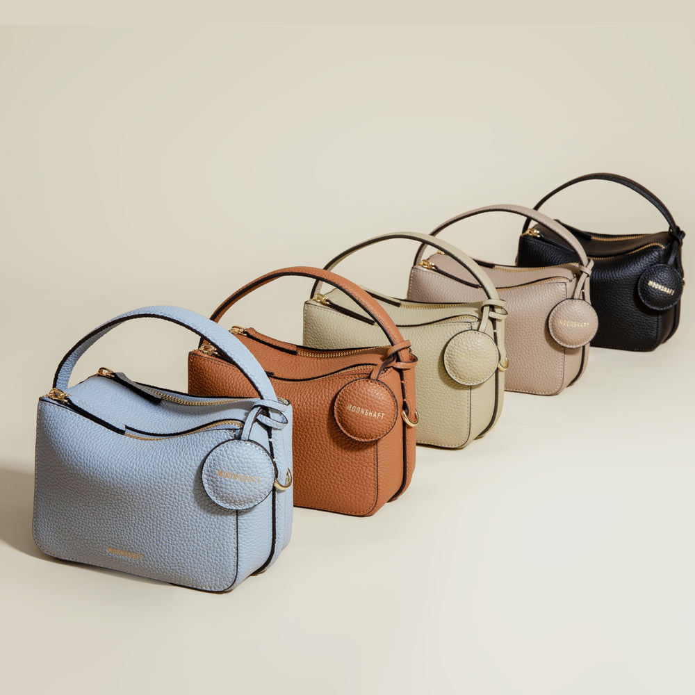 A lineup of the Mini Double Zip Bags in five different colors: aquamarine, toffee, beige, taupe, and black. Each bag is made from pebbled leather and features a round leather tag embossed with the 'MOONSHAFT' logo in gold. The structured design, gold-tone hardware, and top handle are consistent across all colors, showcasing the versatility and elegant style of the collection.