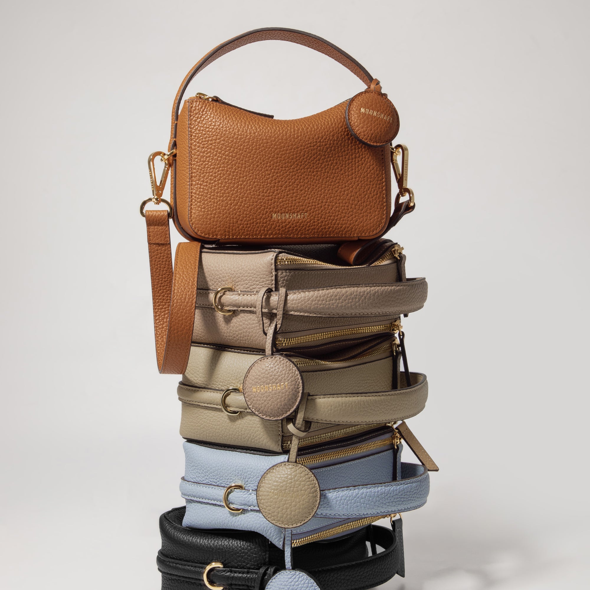 A stacked display of Mini Double Zip Bags in five colors: toffee, taupe, beige, aquamarine, and black. Each bag, made from pebbled leather, features gold-tone hardware, a top handle, and a round leather tag embossed with the 'MOONSHAFT' logo. The structured, minimalist design of the bags is highlighted in this vertical arrangement, showcasing their elegant and versatile style