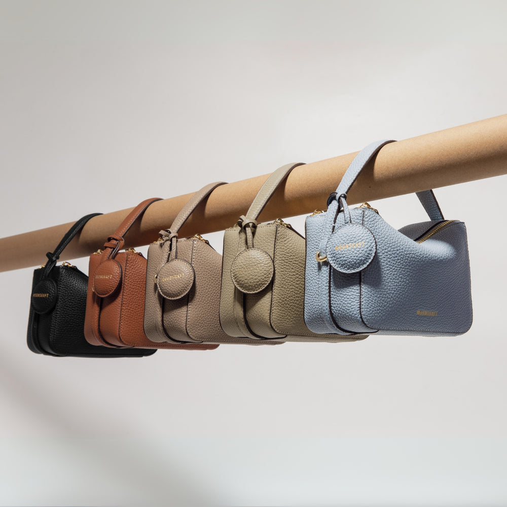 A collection of Mini Double Zip Bags hanging on a display rod, featuring various colors: black, toffee, taupe, beige, and aquamarine. Each bag is made from pebbled leather and features a top handle, gold-tone hardware, and a round leather tag embossed with the 'MOONSHAFT' logo. The sleek, structured design and color variety showcase the versatility and timeless elegance of the collection.