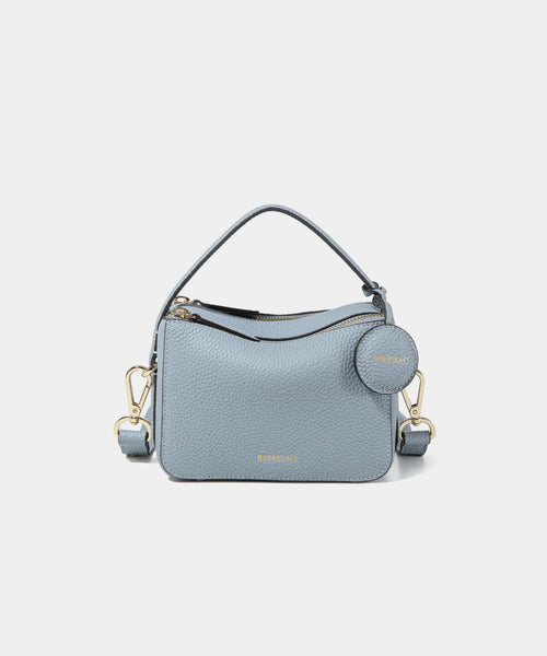 A close-up of the Mini Double Zip Bag in a soft blue shade, made from pebbled leather. The bag features a top handle and an adjustable shoulder strap with gold-tone hardware. It has two zippered compartments for storage and a round leather tag attached to the side, embossed with the brand logo 'MOONSHAFT' in gold. The minimalist and structured design enhances its modern and stylish appeal.