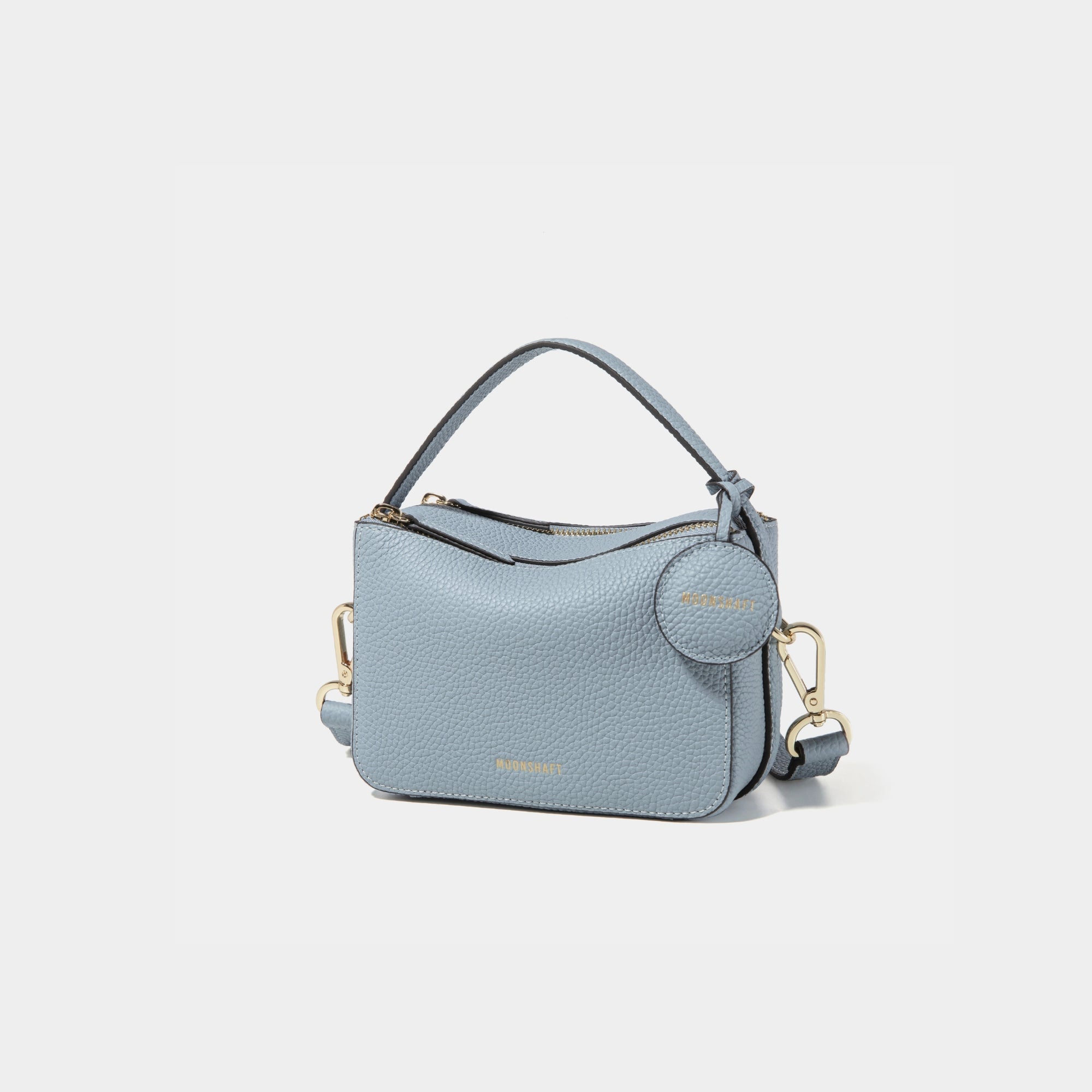A side-front view of the Mini Double Zip Bag in aquamarine color, made from pebbled leather. The bag features a top handle, gold-tone hardware, and an adjustable shoulder strap. It has two zippered compartments and a small round leather tag attached to the side, embossed with the 'MOONSHAFT' logo in gold. The clean and structured design showcases its minimalist elegance and functional style.