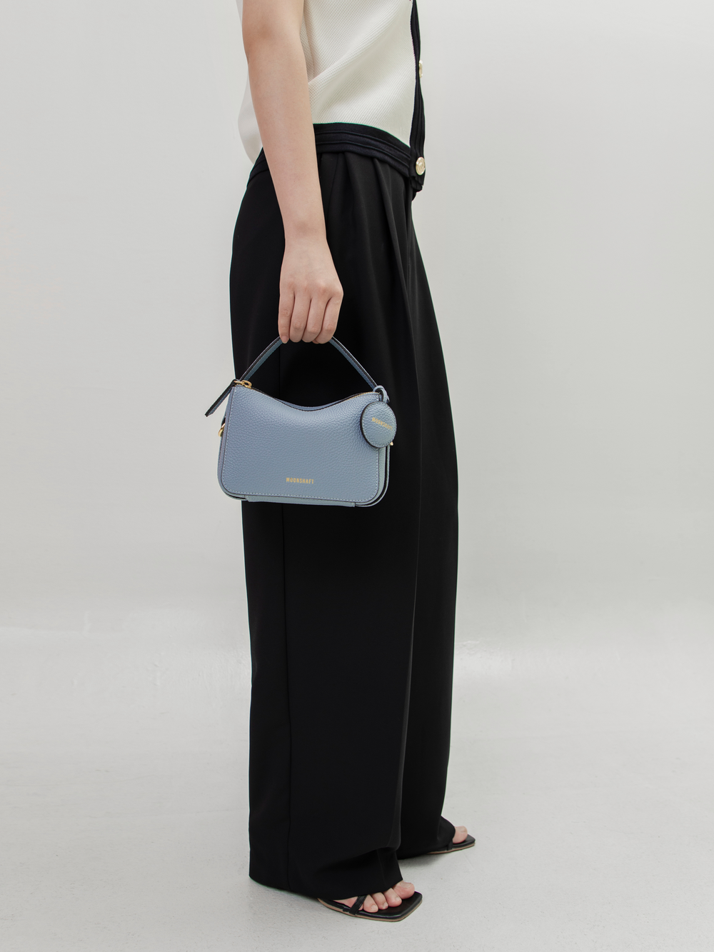 A model dressed in black wide-leg trousers and sandals carries the Mini Double Zip Bag in aquamarine by its top handle. The bag, made from pebbled leather, features gold-tone hardware and a round leather tag embossed with the 'MOONSHAFT' logo. The minimalist and structured design of the bag complements the model’s chic, modern outfit.