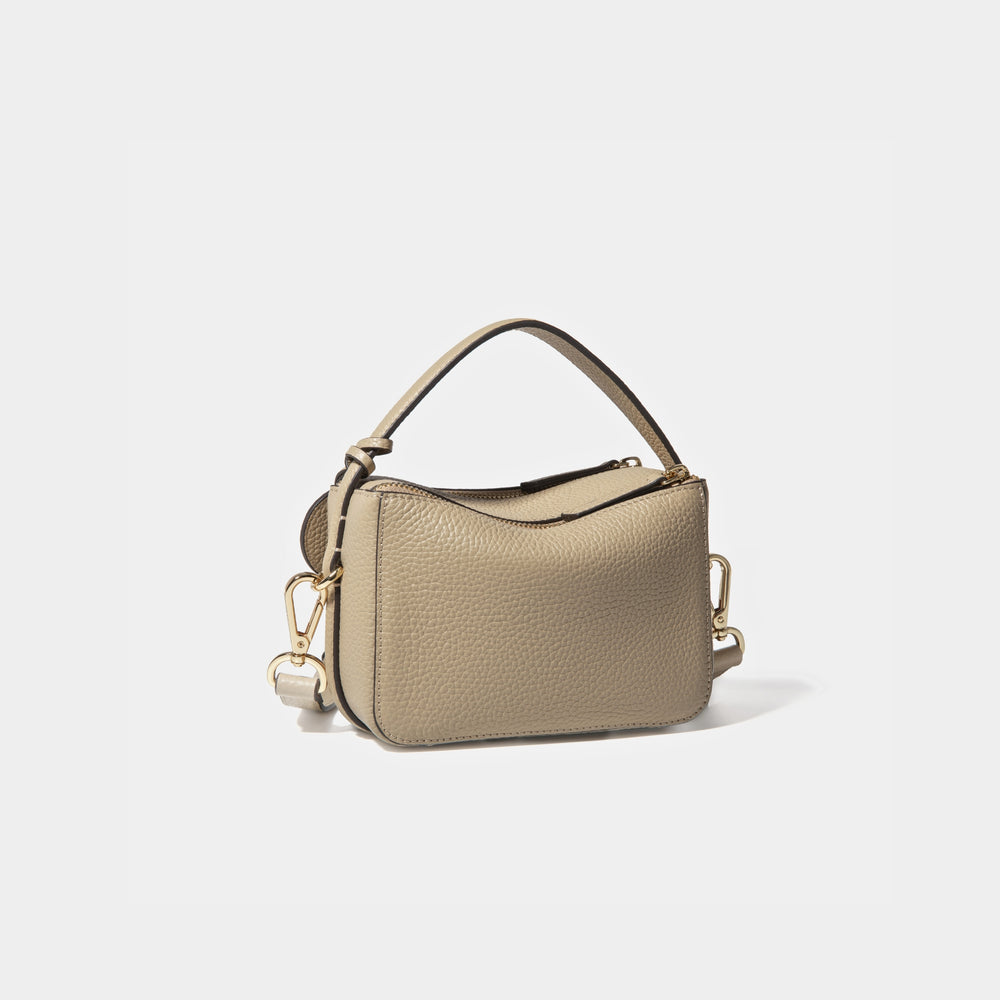 A diagonal back view of the Mini Double Zip Bag in beige color, crafted from pebbled leather. The bag features a smooth and structured design with a top handle and detachable gold-tone hardware. The clean back panel showcases the minimalist style, while the adjustable shoulder strap and two zippered compartments add both functionality and elegance to the overall look.