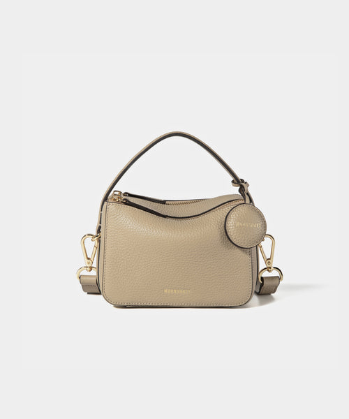 A close-up of the Mini Double Zip Bag in beige color, made from pebbled leather. The bag features a top handle and an adjustable shoulder strap with gold-tone hardware. It has two zippered compartments for storage and a round leather tag attached to the side, embossed with the brand logo 'MOONSHAFT' in gold. The minimalist and structured design enhances its modern and stylish appeal.